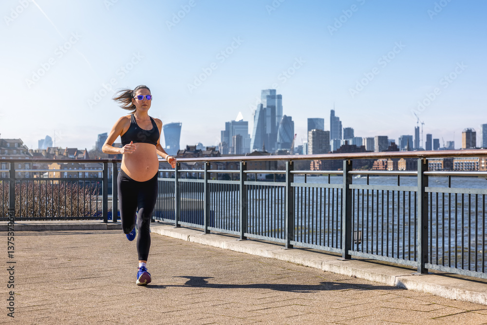 A athletic, pregnant woman in sports clothing running for exercise in front of an urban skyline during a sunny day