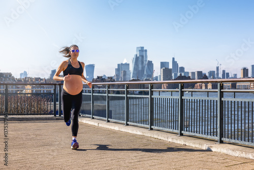 Obraz na płótnie A athletic, pregnant woman in sports clothing running for exercise in front of an urban skyline during a sunny day w salonie