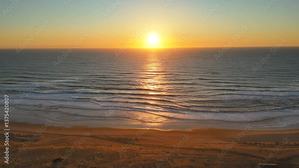 Aerial drone shot of golden sunset over calm ocean, with waves reflecting light and a peaceful sandy beach in the foreground view. A tranquil, scenic coastal moment at dusk. Aerial drone shot of golden sunset over calm ocean, with waves reflecting light and a peaceful sandy beach in the foreground view. A tranquil, scenic coastal moment at dusk.