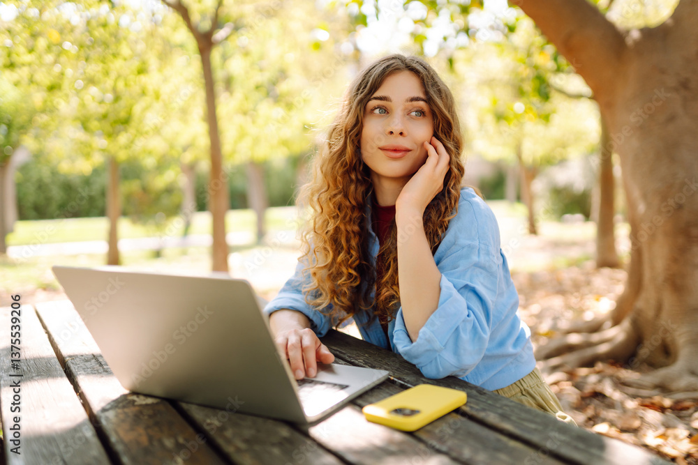Young woman working at a table in the park on a laptop. Happy female freelancer enjoying the weather outdoors. Remote work concept. Lifestyle. Young woman working at a table in the park on a laptop. Happy female freelancer enjoying the weather outdoors. Remote work concept. Lifestyle.