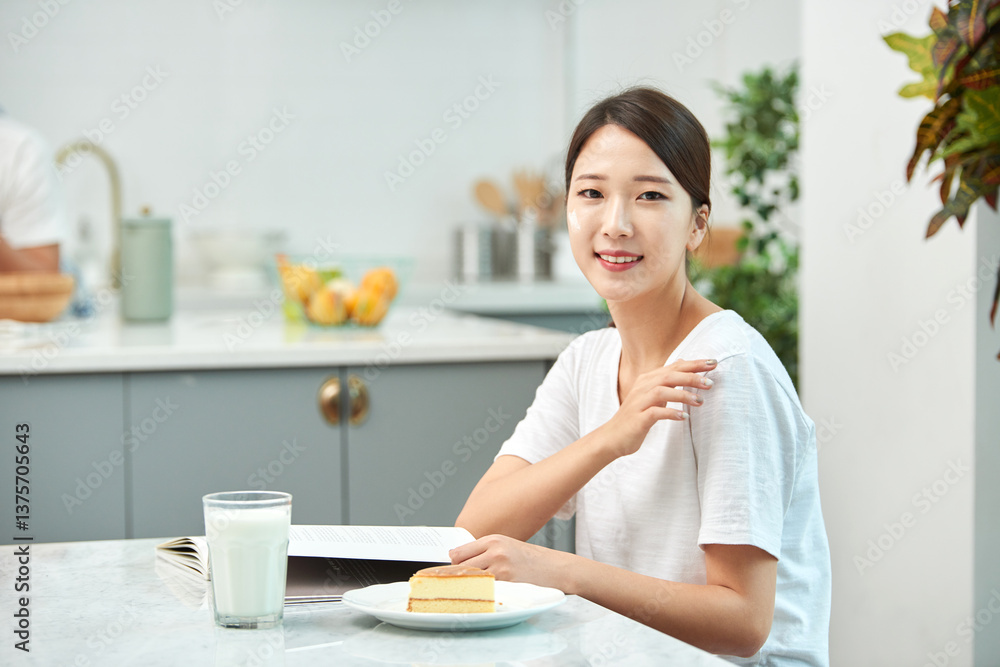Young Asian woman in a white T-shirt sitting at a kitchen dining table chair, reading while eating bread and drinking milk Young Asian woman in a white T-shirt sitting at a kitchen dining table chair, reading while eating bread and drinking milk