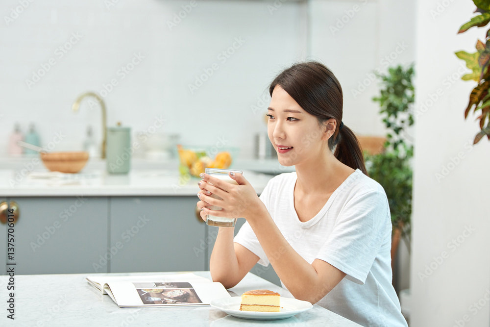 Young Asian woman in a white T-shirt sitting at a kitchen dining table chair, reading while eating bread and drinking milk Young Asian woman in a white T-shirt sitting at a kitchen dining table chair, reading while eating bread and drinking milk