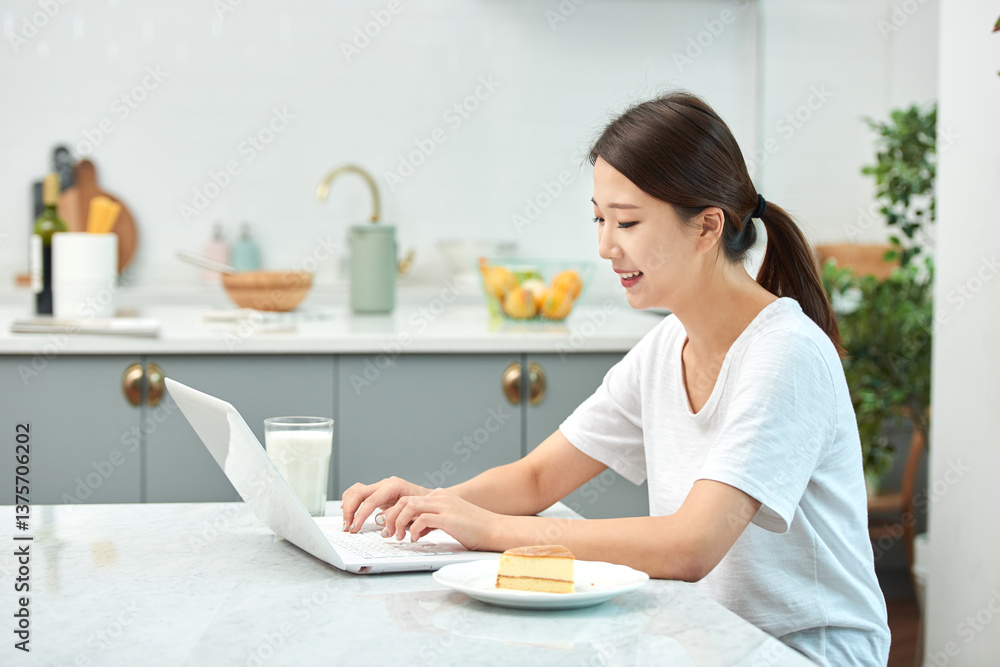 Young Asian woman in a white T-shirt sitting at a kitchen dining table chair, working on a laptop while eating bread and drinking milk Young Asian woman in a white T-shirt sitting at a kitchen dining table chair, working on a laptop while eating bread and drinking milk