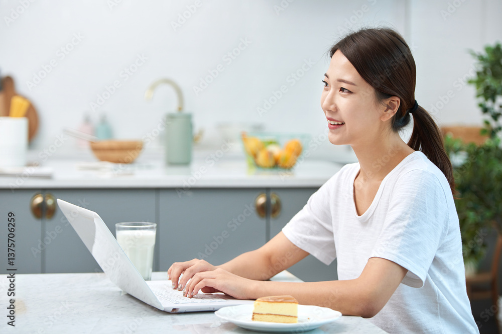 Young Asian woman in a white T-shirt sitting at a kitchen dining table chair, working on a laptop while eating bread and drinking milk Young Asian woman in a white T-shirt sitting at a kitchen dining table chair, working on a laptop while eating bread and drinking milk
