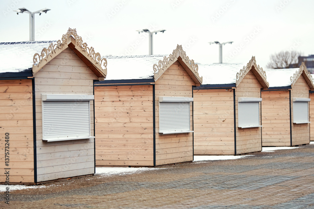 Shuttered wooden kiosks in an outdoor market area, arranged in perfect row, with snow covered rooftops, waiting for the next seasonal event. Holiday market in a city square. Selective focus