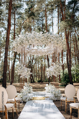 A luxurious wedding ceremony in a pine forest with elegant white chairs featuring mirrored legs, a tall arch adorned with white flowers, and a pristine white aisle leading to the altar