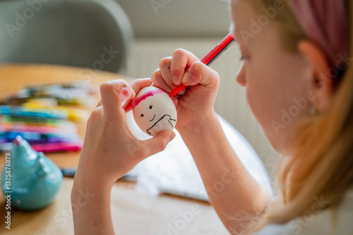 girl 8 years old blonde sits on a chair at home and paints a chicken white egg for the holy Orthodox holiday of bright Easter in the kitchen at home, bright family photo