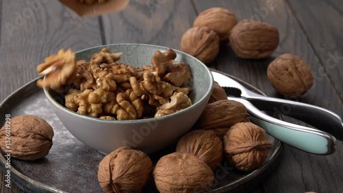 Bowl of walnuts being poured into it from unseen container surrounded by whole walnuts on wooden table.