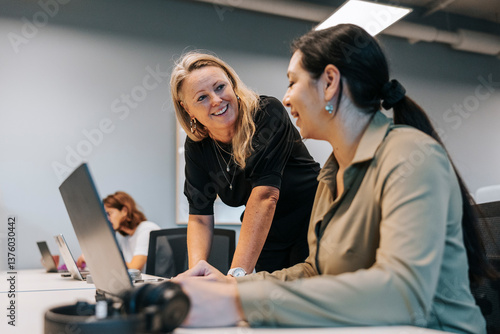 Happy blond businesswoman talking with coworker sitting at desk in office