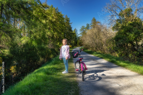 A cheerful and lively young girl happily stands beside her bicycle on a beautiful picturesque trail, enjoying a delightful sunny day in a lush green landscape, Bohernabreena Reservoir, Dublin, Ireland