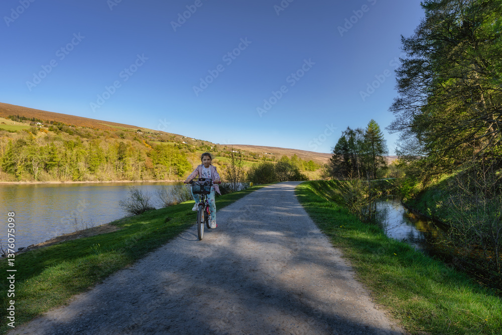 A young girl joyfully rides her bicycle along beautiful, picturesque river pathway which is surrounded by vibrant autumn foliage, creating a serene atmosphere, Bohernabreena Reservoir, Dublin, Ireland