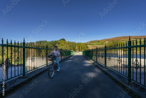 Obraz na płótnie A child joyfully rides a colorful bicycle across a picturesque bridge, amidst lush greenery and a clear blue sky, perfect for outdoor adventures and freedom, Bohernabreena Reservoir, Dublin, Ireland w salonie