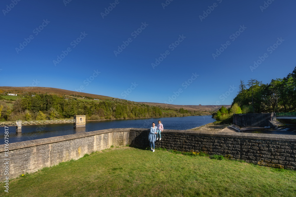 A serene landscape showcases a mother and daughter on s tranquil dam and reservoir, surrounded by trees, all set beneath a clear blue sky on a bright day, Bohernabreena Reservoir, Dublin, Ireland