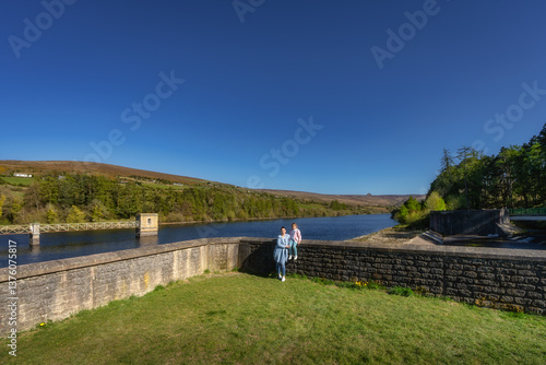 Obraz na płótnie A serene landscape showcases a mother and daughter on s tranquil dam and reservoir, surrounded by trees, all set beneath a clear blue sky on a bright day, Bohernabreena Reservoir, Dublin, Ireland w salonie