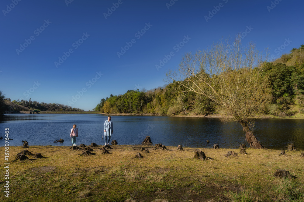 A picturesque lakeside scene that features mother and daughter standing on tree stumps by the waters edge entirely surrounded by beautiful nature and greenery, Bohernabreena Reservoir, Dublin, Ireland