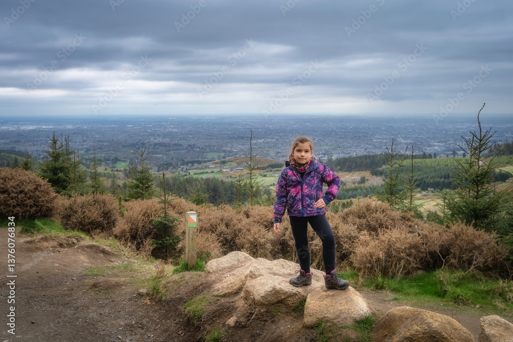 A young girl stands confidently on a large rock formation, savoring the breathtaking views of the expansive horizon from a lush, scenic mountain trail nearby, Wicklow, Dublin, Ireland