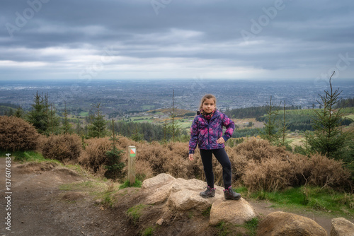 Obraz na płótnie A young girl stands confidently on a large rock formation, savoring the breathtaking views of the expansive horizon from a lush, scenic mountain trail nearby, Wicklow, Dublin, Ireland w salonie