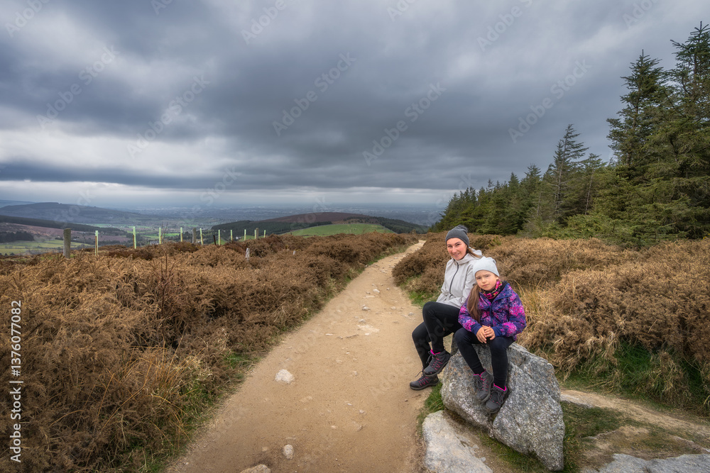 Two enthusiastic hikers pause for a break on a large stone, taking in the breathtaking views and the natural tranquility that surrounds them under a cloudy sky, Wicklow, Dublin, Ireland