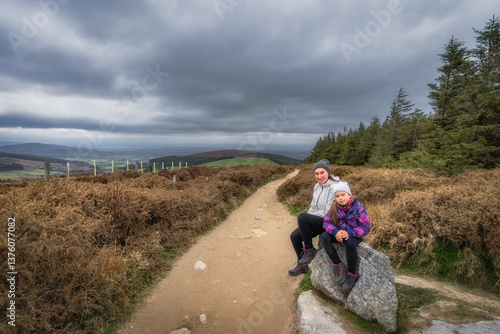 Obraz na płótnie Two enthusiastic hikers pause for a break on a large stone, taking in the breathtaking views and the natural tranquility that surrounds them under a cloudy sky, Wicklow, Dublin, Ireland w salonie