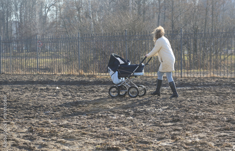 A young mother in a white down jacket with a baby stroller walks through the spring land, Tovarishchesky Prospect, Saint Petersburg, Russia, April 01, 2025 A young mother in a white down jacket with a baby stroller walks through the spring land, Tovarishchesky Prospect, Saint Petersburg, Russia, April 01, 2025