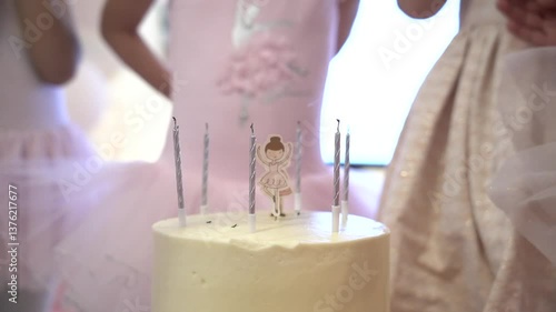 A close-up of a white birthday cake with silver candles and a ballerina topper, surrounded by children in pastel party dresses. Perfect for ballet-themed celebrations and childhood moments.

