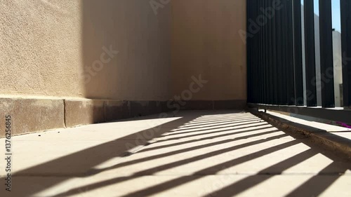 Sunlit outdoor corridor with strong geometric shadows from a metal fence. Warm beige walls and tiled flooring create a minimalist, modern aesthetic. Ideal for architectural and abstract concepts.