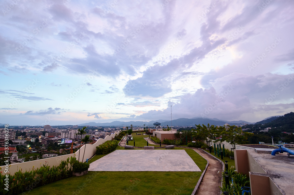 Fototapeta premium A scenic rooftop garden at sunset with a view of a city skyline and distant mountains under a cloudy sky.