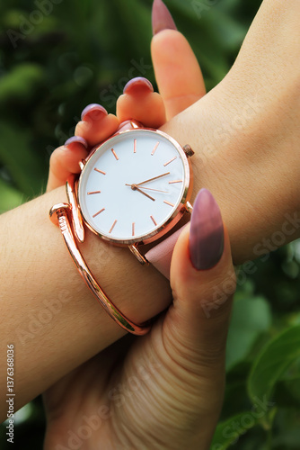 Close-up of wrist adorned with hand with long, manicured nails adjusts minimalist watch, and rose gold bracelet against lush green background, showcasing elegance and style in natural setting.