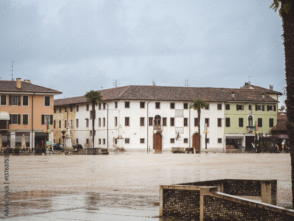Obraz premium A rainy day in the historic town square of Palmanova, Italy, featuring old buildings with colorful facades, statues, and palm trees under a cloudy sky.