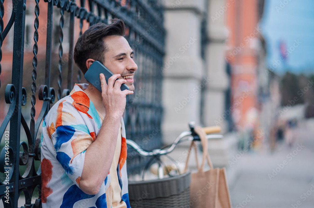 Fototapeta premium Young man talking on smartphone leaning on a gate with his bicycle nearby