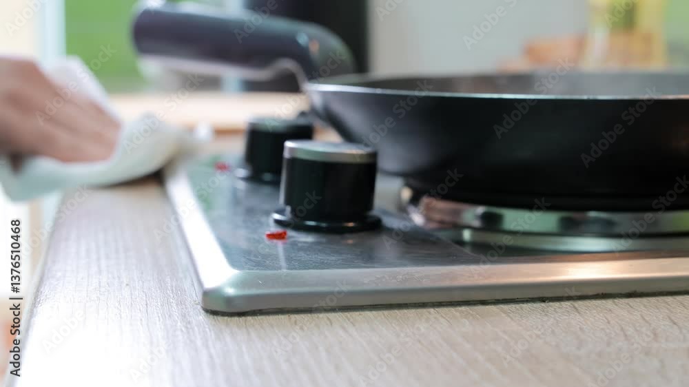 A hand wipes the stove surface near control knobs and a frying pan using a paper towel, showing kitchen cleanliness habits during cooking process A hand wipes the stove surface near control knobs and a frying pan using a paper towel, showing kitchen cleanliness habits during cooking process