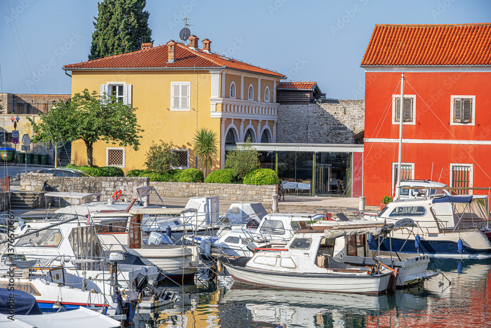 A picturesque Mediterranean harbor with colorful historic buildings, moored boats, and a tranquil waterfront, blending old-world charm with modern elements under a bright blue sky.