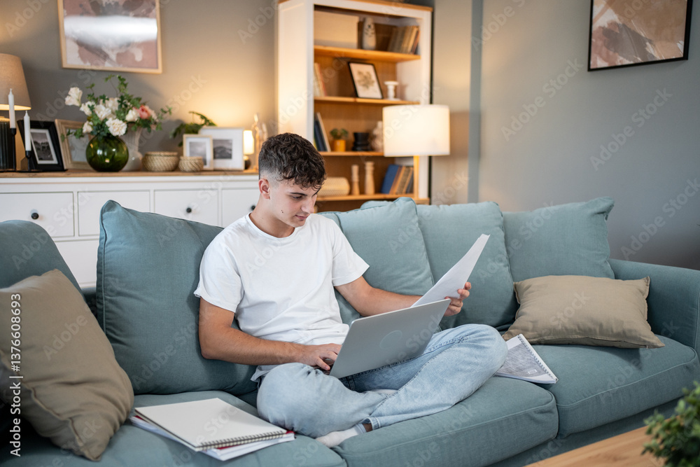 Focused student studying online and reviewing documents on sofa at home Focused student studying online and reviewing documents on sofa at home