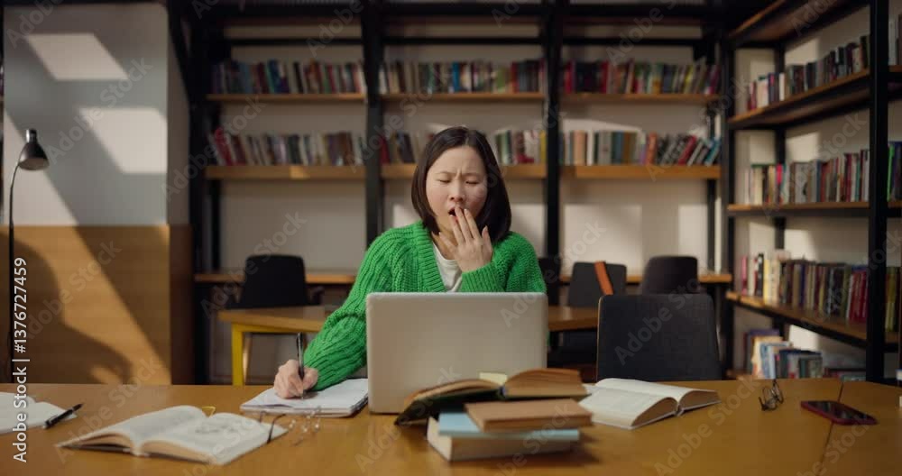 Sleepy girl student in a green sweater makes notes in a notebook and falls asleep in front of a laptop in the library Sleepy girl student in a green sweater makes notes in a notebook and falls asleep in front of a laptop in the library