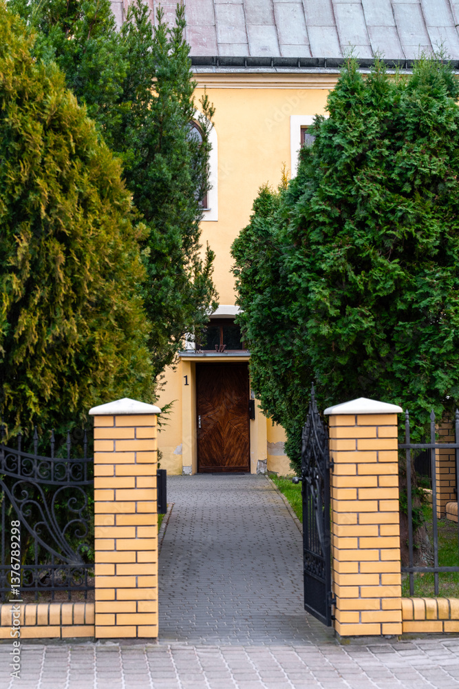 Naklejka premium Wejście do posesji z otwartą bramą i drzewami – Przestrzeń miejska z zielenią w tle.