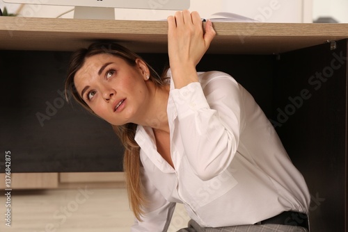 Scared young woman hiding under office desk during earthquake