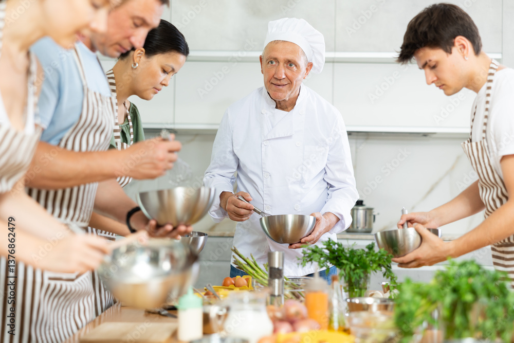 Elderly man cook in uniform teaches group of different people how to cook dish Elderly man cook in uniform teaches group of different people how to cook dish