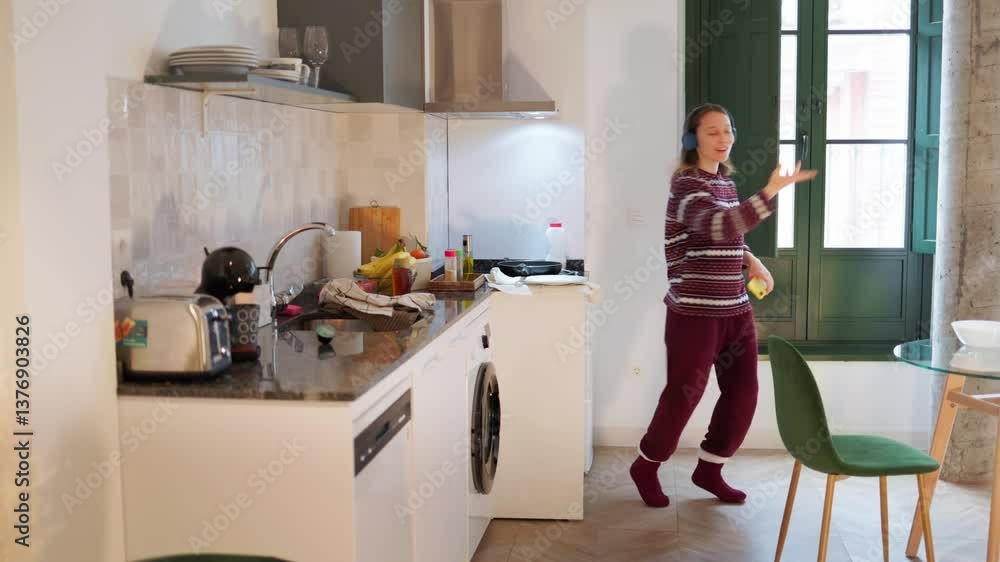 Happy woman wearing pajamas and headphones, dancing and listening to music while preparing breakfast in kitchen Happy woman wearing pajamas and headphones, dancing and listening to music while preparing breakfast in kitchen