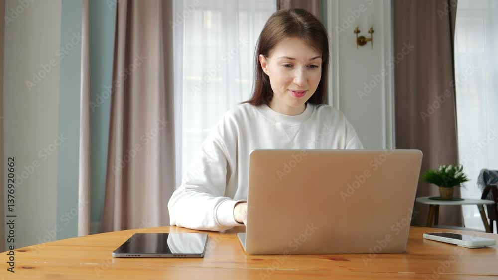 Focused individual uses a laptop while seated at a wooden table in a bright living space. Focused individual uses a laptop while seated at a wooden table in a bright living space.