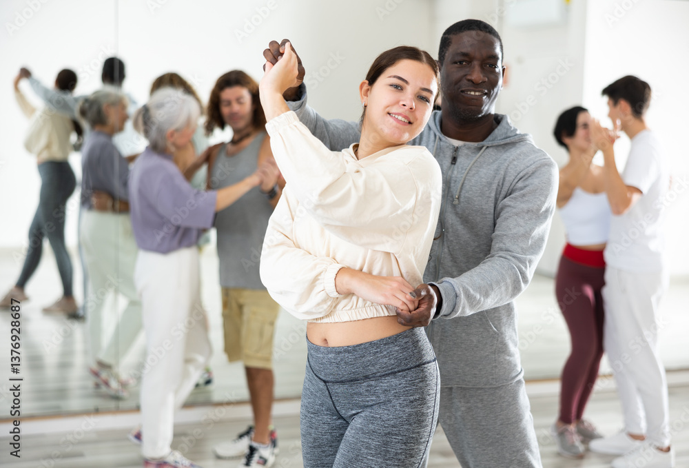 Naklejka premium Young smiling girl learning to dance waltz paired with african american man in dancing class..
