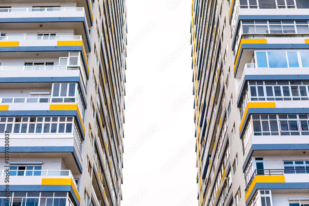 Two contemporary apartment buildings showcasing a blend of white, blue, and yellow facades. Parallel positioning creates a narrow space between them, leading to bright overexposed sky in background