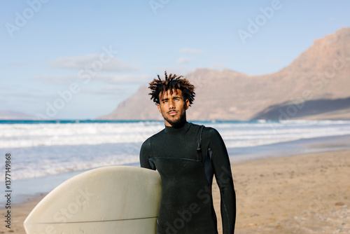 Black man wearing a neoprene wetsuit, posing with his surfboard