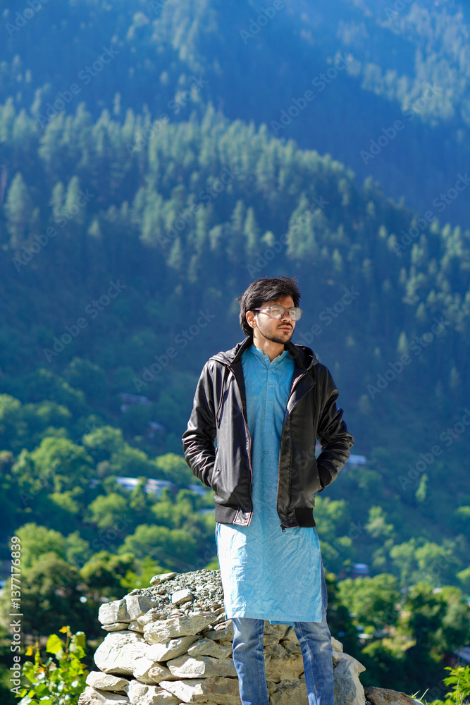Naklejka premium Young traveler in a black jacket and blue jeans stands on rocks with hands in pockets, forested mountains provide a serene backdrop.