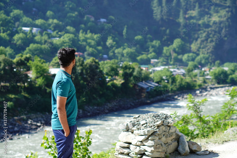Naklejka premium A young black male stands outdoors, gazing at a green valley with a river. Scenic nature and peace surround them.