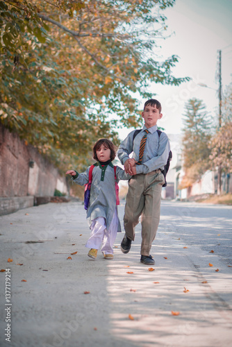 Elder brother holding hand of younger sister, in school uniform and backpacks, children walking to school.