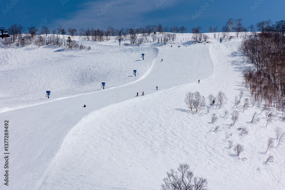 Snowboarders descending a wide, steep ski slope on bright sunny day