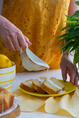 Woman in yellow clothes pouring cream on lemon cake.
