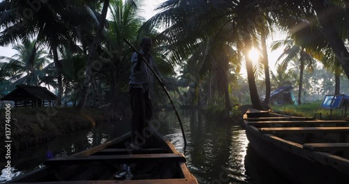 Silhouette of canoe driver gently pushing through Kerala backwaters at sunrise. Traditional boat glides under palm trees in warm light, evoking peaceful travel and cultural authenticity.