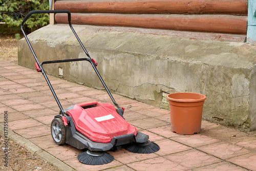 Red outdoor power sweeper rests on a paved surface with black bristles visible, accompanied by an orange bucket, set against a wooden and stone building exterior.