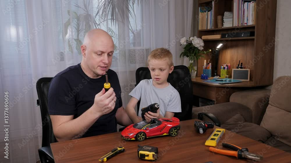 A man and a little boy are sitting together at a table and repairing a broken car with a screwdriver. Young caucasian father helps his son fix his favorite toy at home.